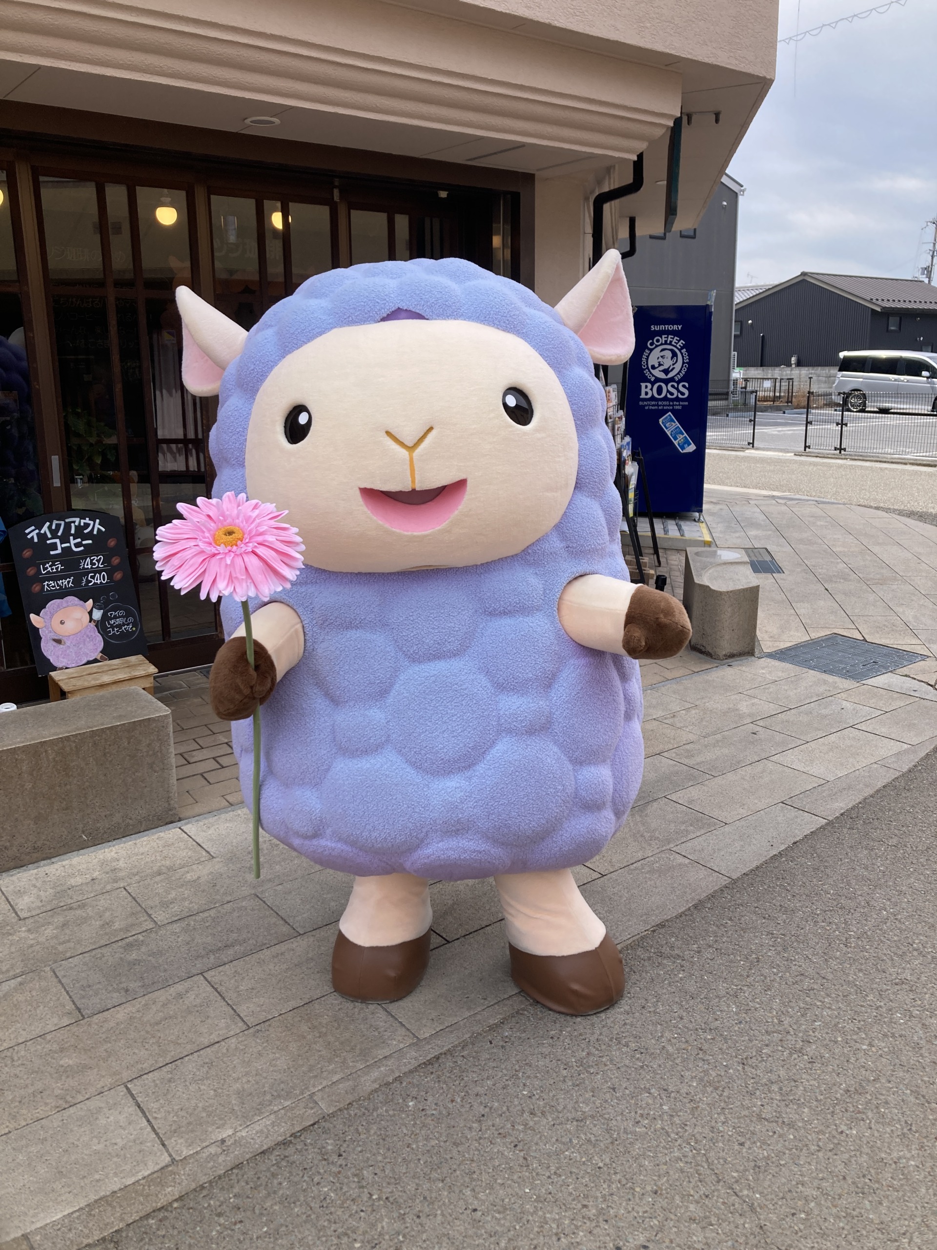 紫の羊の着ぐるみがピンクのガーベラを持って、店の入口の前に立っている全身